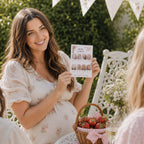A smiling pregnant woman in a white floral dress holds up a Berry Sweet Baby Bump or Beer picture quiz card during an outdoor strawberry themed baby girl baby shower with friends.. A wicker basket of fresh strawberries and a glass of champagne sit on the table next to her, with white bunting and green foliage in the background.