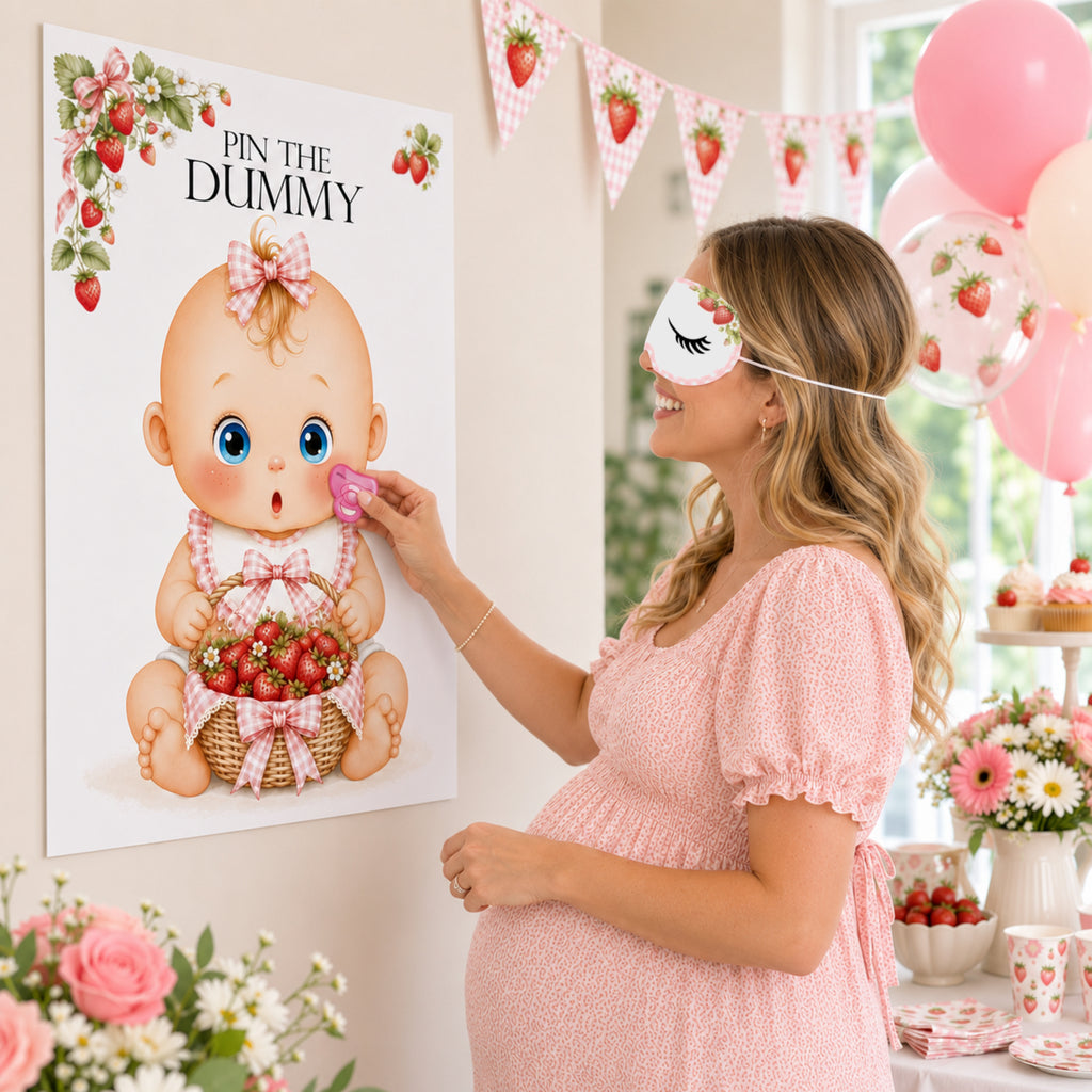 A pregnant woman in a pink dress and white sleep mask participates in a baby shower game, pinning a pink teething toy to a large wall poster that reads 'PIN THE DUMMY' with an illustration of a baby holding strawberries. The scene is decorated with pink balloons, strawberry bunting, and a dessert table.