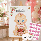 A baby shower game board or sign on an easel that reads 'PIN THE DUMMY,' featuring a cartoon baby holding strawberries. A sheet of pink pacifier stickers and a matching sleep mask are placed beside it, with a softly blurred party background of balloons, flowers, and bunting.