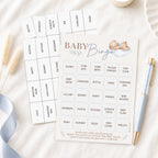 Baby Bingo party game cards laid out on a soft white fabric surface with a light blue ribbon, a pen, and a small candle. The front card shows the title 'BABY Bingo' with a baby illustration, and the back card lists baby-themed words such as bottle, shower, nursery, and twins.