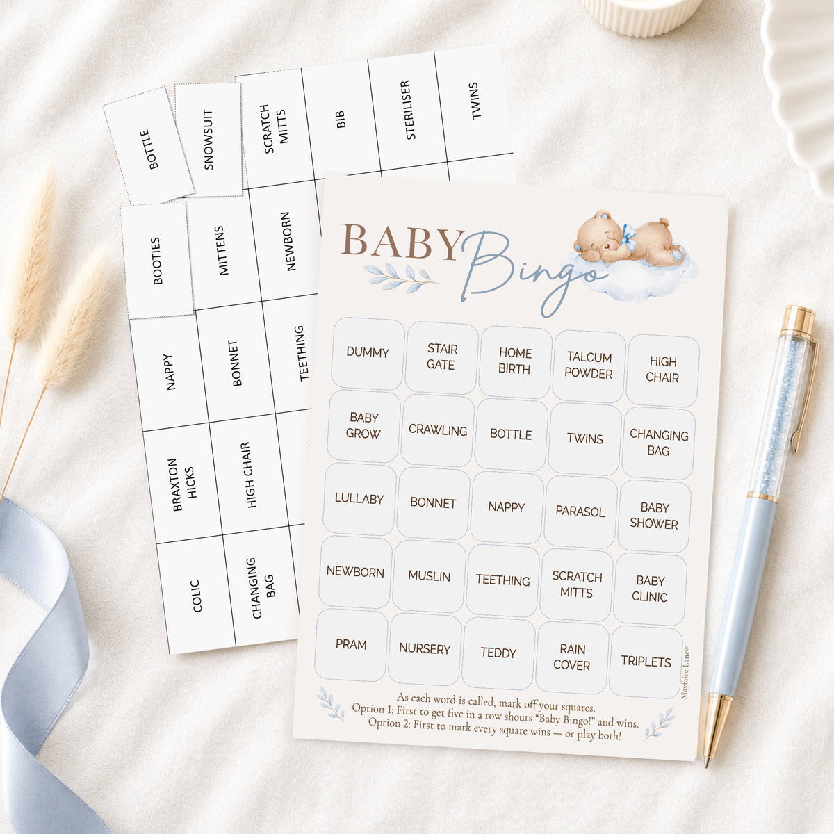 Baby Bingo party game cards laid out on a soft white fabric surface with a light blue ribbon, a pen, and a small candle. The front card shows the title 'BABY Bingo' with a baby illustration, and the back card lists baby-themed words such as bottle, shower, nursery, and twins.