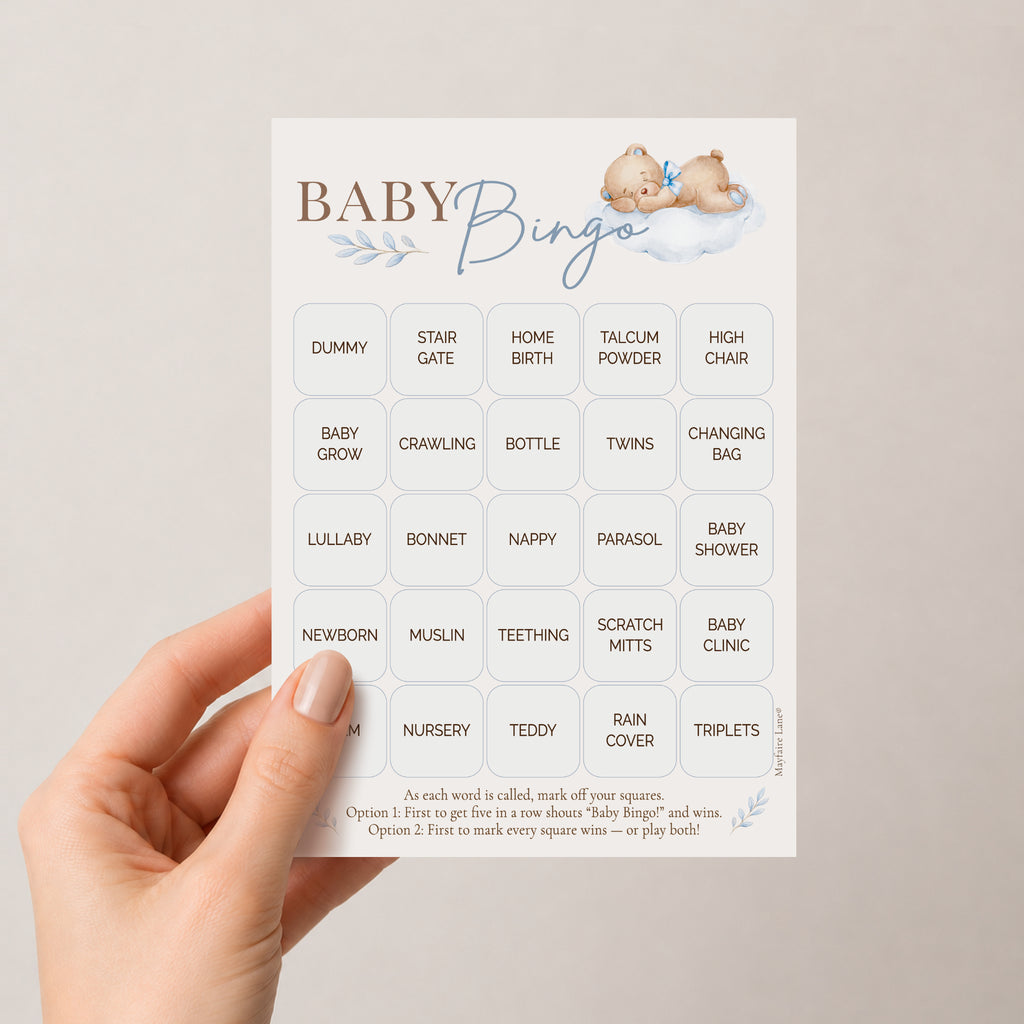 A hand holds a baby bingo card with cute illustrations of two teddy bears on a soft cloud. The card features various baby-themed words in a grid with a playful font and a light blue decorative leaf motif.