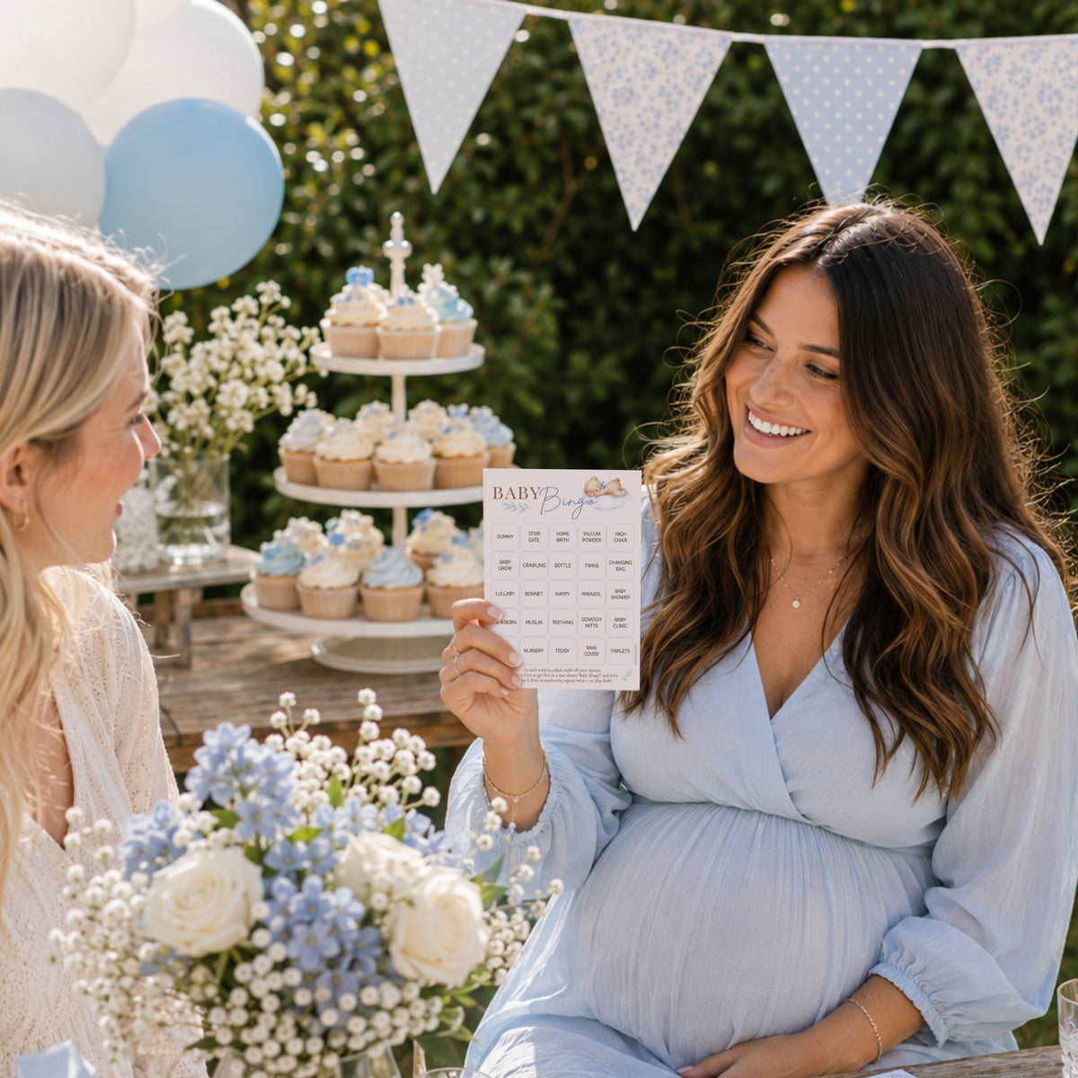 A pregnant woman in a light blue dress holds a 'Baby Shower Bingo' game card at an outdoor baby shower. Another woman is seated beside her, smiling at the game.