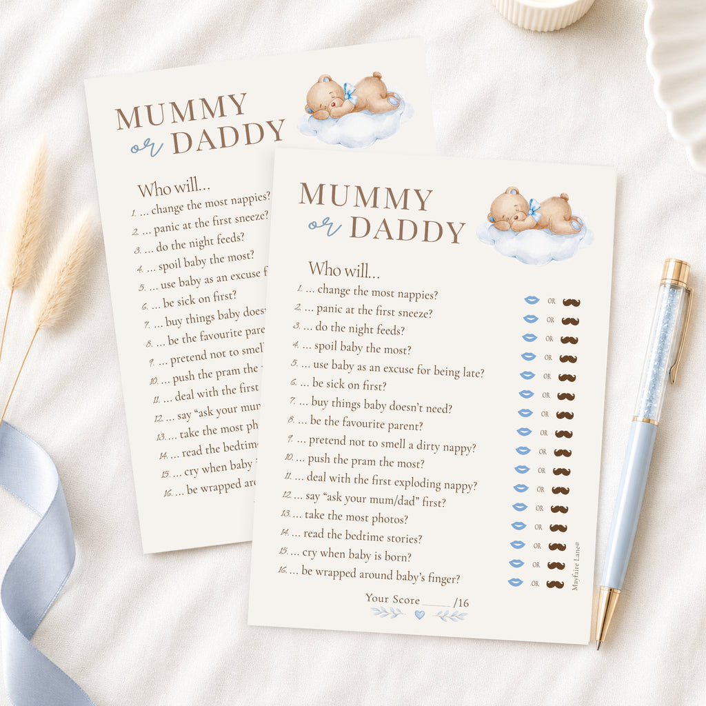 A baby shower game card titled 'Mummy or Daddy' with printed text and small illustrations of a baby and clouds, styled on a white surface with a blue ribbon, a gold pen, and a small cup.