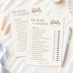 A baby shower game card titled 'Mummy or Daddy' with printed text and small illustrations of a baby and clouds, styled on a white surface with a blue ribbon, a gold pen, and a small cup.