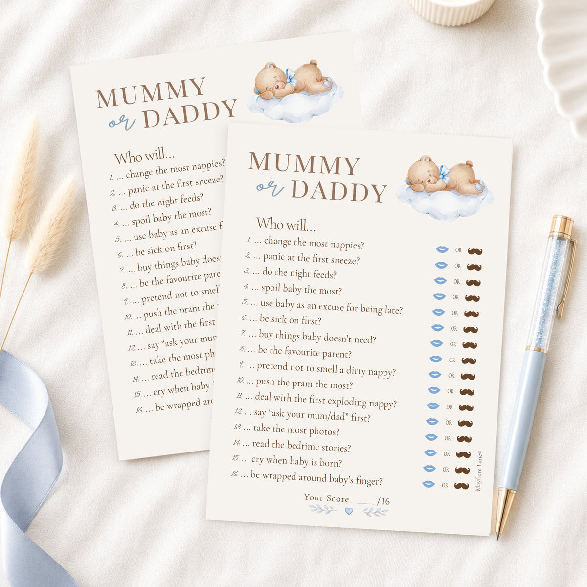 A baby shower game card titled 'Mummy or Daddy' with printed text and small illustrations of a baby and clouds, styled on a white surface with a blue ribbon, a gold pen, and a small cup.