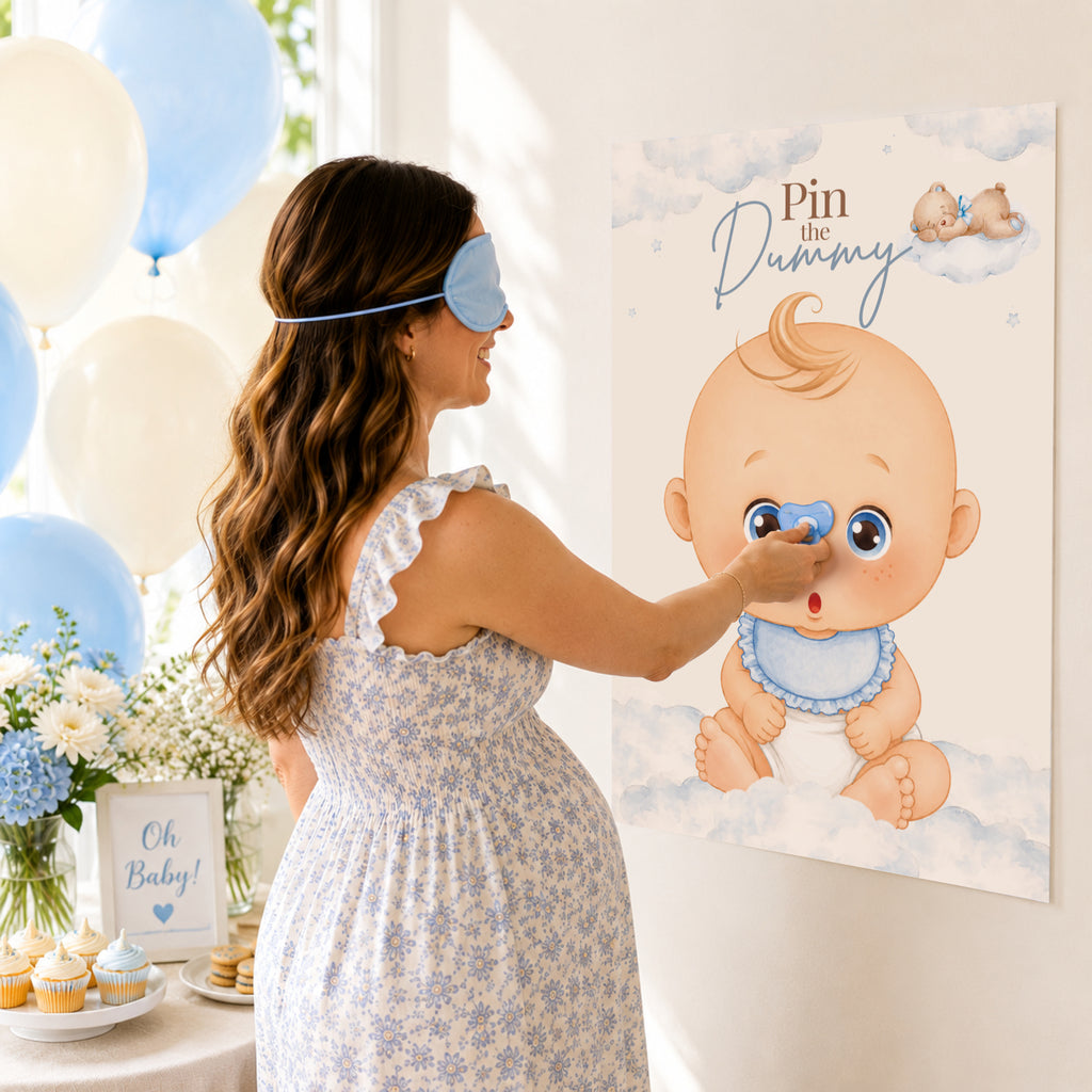 A pregnant woman wearing a blue sleep mask and a light blue floral dress pins a baby-themed canvas print that reads 'Pin the Dummy' on a wall in a decorated baby shower setting with balloons and cupcakes.