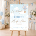 A personalized baby shower sign with a hot air balloon sky theme, displayed on a wooden easel in a decorated party setting with balloons and bunting.