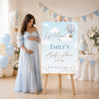A pregnant woman in a light blue off-the-shoulder maternity gown stands beside a personalized baby shower sign on a wooden easel in a decorated baby shower room with balloons, bunting, and a cake table.