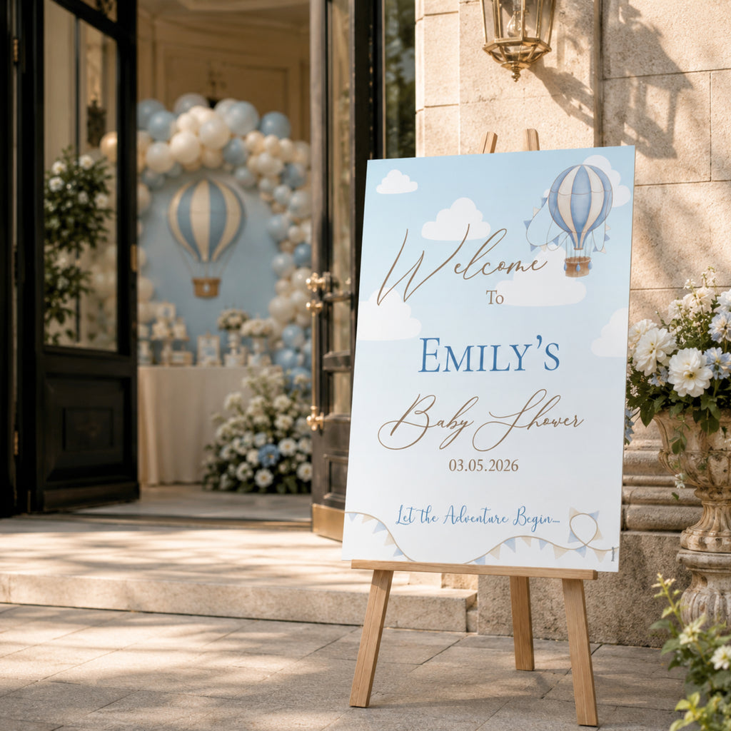 A personalized baby shower welcome sign on a wooden easel, featuring a light blue sky and hot air balloon theme, set in an outdoor event venue with stone walls and floral arrangements.