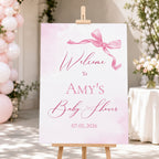 A personalized baby shower welcome sign on a wooden easel, featuring a pink watercolor background, a pink ribbon graphic, and the text 'Welcome to Amy's Baby Shower' with the date '07.05.