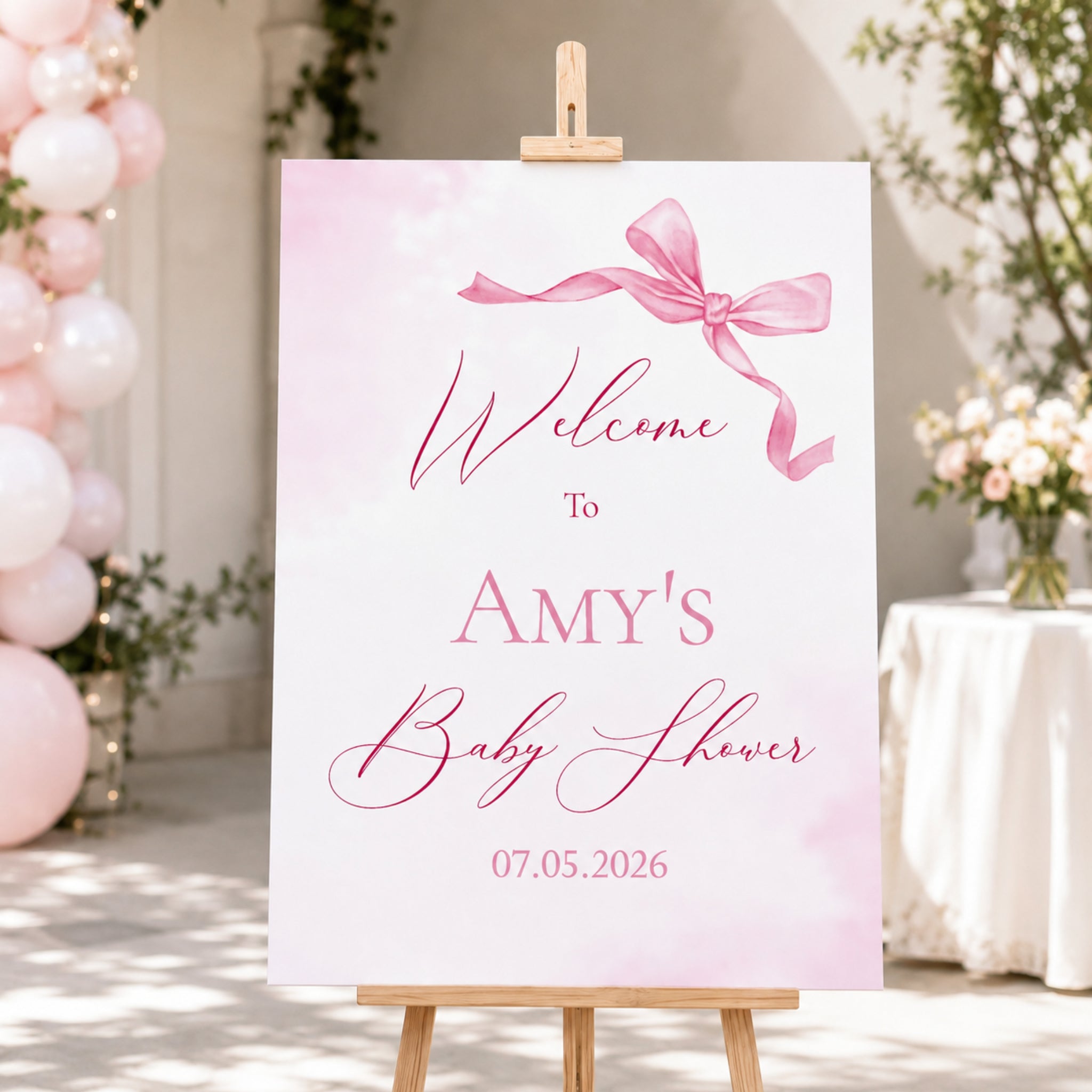 A personalized baby shower welcome sign on a wooden easel, featuring a pink watercolor background, a pink ribbon graphic, and the text 'Welcome to Amy's Baby Shower' with the date '07.05.