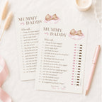 Two printed baby shower game cards titled "Mummy or Daddy" laid on a white surface, with a pink pen, small cup, and dried grass stems around them.
