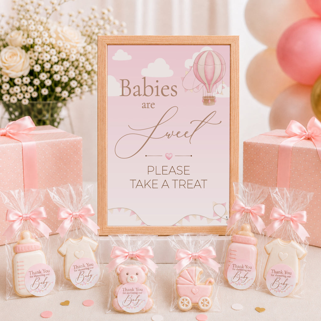 A baby shower themed treat table display featuring a central framed sign that reads 'Babies are Sweet PLEASE TAKE A TREAT' with a hot air balloon illustration. Surrounding the sign are baby bottle-shaped cookies in cellophane bags with pink bows, a teddy bear cookie, a stroller cookie, and pink gift boxes with ribbons.