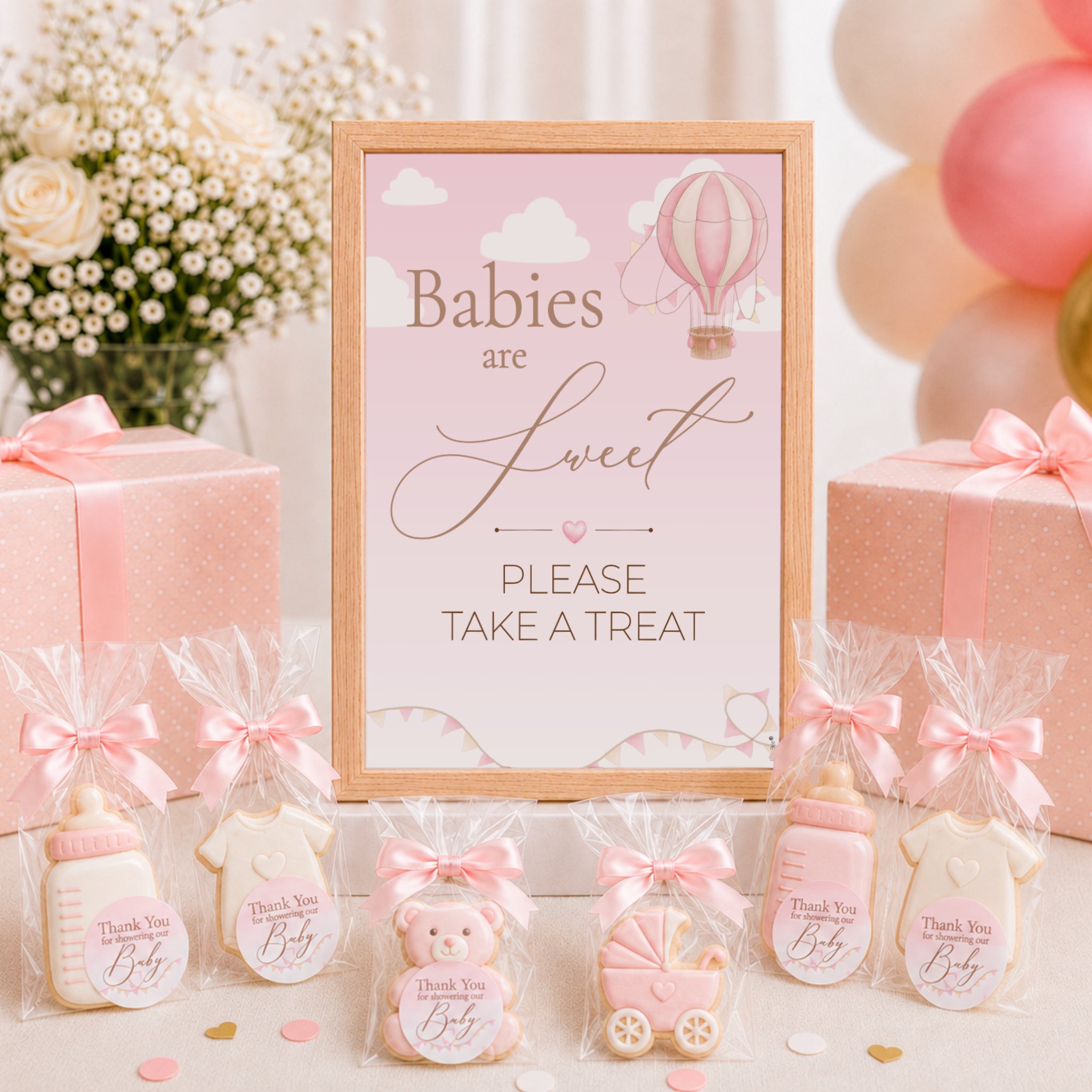A baby shower themed treat table display featuring a central framed sign that reads 'Babies are Sweet PLEASE TAKE A TREAT' with a hot air balloon illustration. Surrounding the sign are baby bottle-shaped cookies in cellophane bags with pink bows, a teddy bear cookie, a stroller cookie, and pink gift boxes with ribbons.