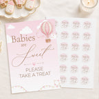A baby treat sign and matching sticker sheet in a soft pink and gold color scheme, styled on a light beige surface with a lit candle and baby-themed cookies in the background.