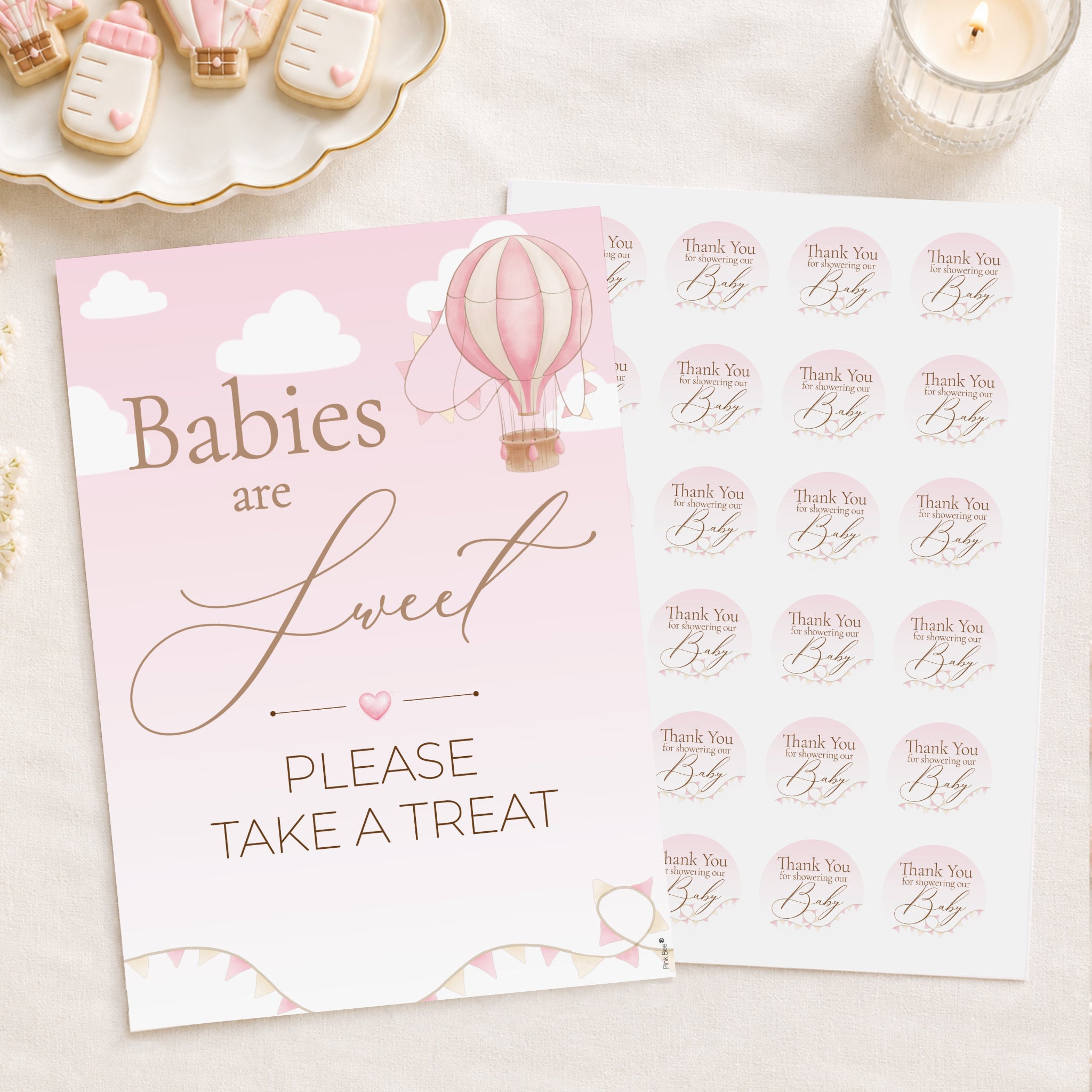 A baby treat sign and matching sticker sheet in a soft pink and gold color scheme, styled on a light beige surface with a lit candle and baby-themed cookies in the background.