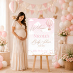 A pregnant woman in a cream maxi dress stands beside a pink and white 'Welcome to Nicole's Baby Shower' sign on a wooden easel in a decorated baby shower room with pink and white balloons, bunting, and floral arrangements.