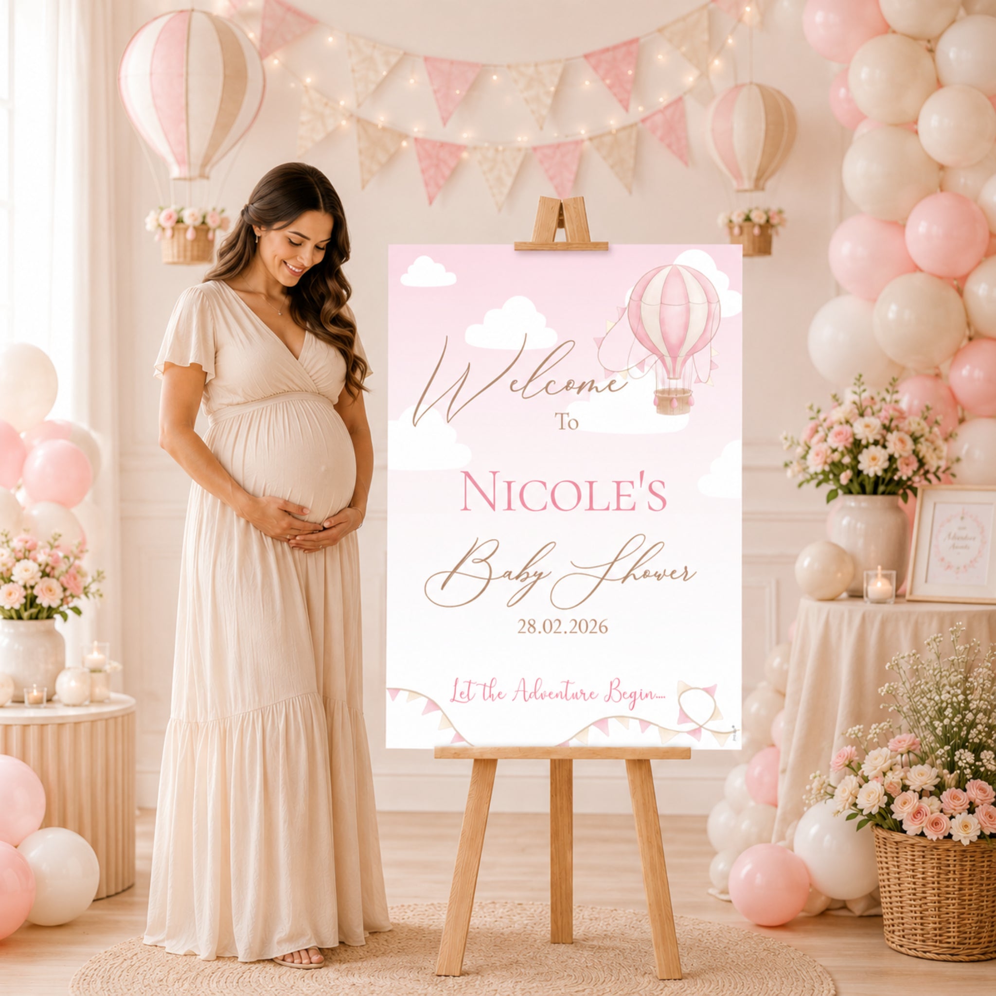 A pregnant woman in a cream maxi dress stands beside a pink and white 'Welcome to Nicole's Baby Shower' sign on a wooden easel in a decorated baby shower room with pink and white balloons, bunting, and floral arrangements.