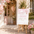 Personalised Up and Away Baby Girl Foamex welcome sign displayed on an easel outside a baby shower venue with pink balloon arch, floral decorations and hot air balloon themed styling.